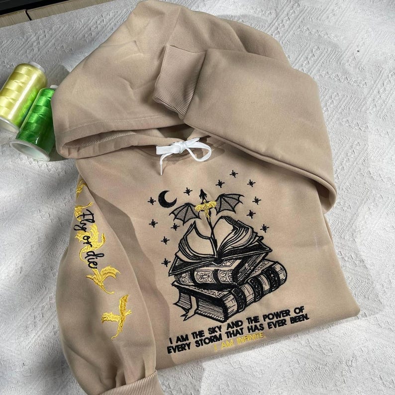 May include: A tan hoodie with a black and gold embroidered design on the front. The design features a stack of books, a crescent moon, a dragon, and stars. The text "I am the sky and the power of every storm that has ever been I am infinite" is embroidered on the front of the hoodie. The text "Fly or die" is embroidered on the left sleeve in gold.