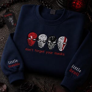 May include: Navy blue sweatshirt with embroidered mask designs in red, white, and black. The text "don't forget your masks" is embroidered in red. Sleeves feature "little trouble" and "little devil" in red.