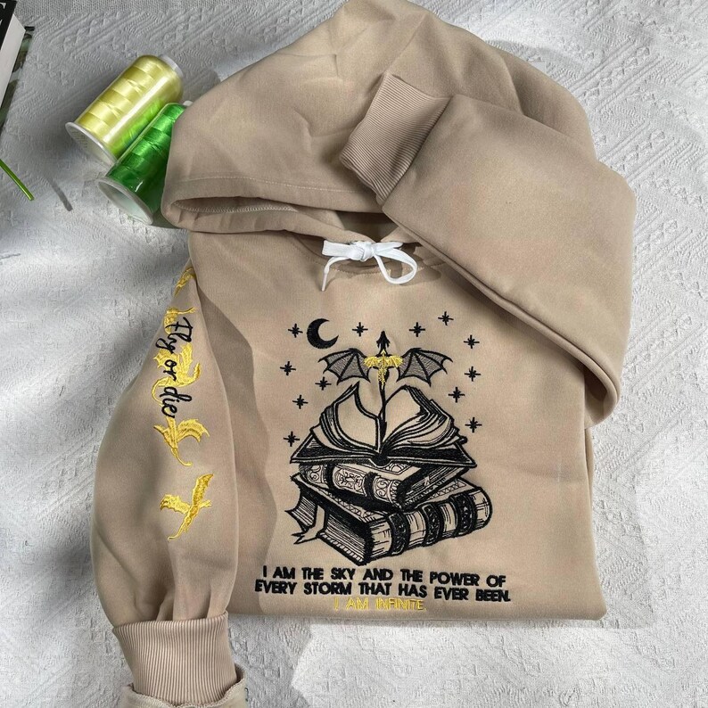 May include: A beige hoodie with a black and yellow embroidered design of a bat, a book, and a crescent moon. The text "Fly or die" is embroidered on the left sleeve. The text "I am the sky and the power of every storm that has ever been. I am infinite." is embroidered on the front.