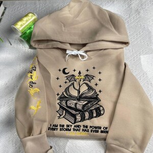 May include: A beige hooded sweatshirt with a black embroidered design of a book with wings and stars. The text "Fly or die" is embroidered on the sleeve and the text "I am the sky and the power of every storm that has ever been I am definite" is embroidered on the front.