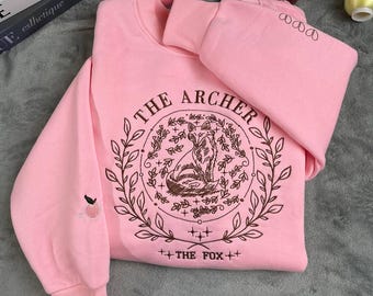 May include: Pink sweatshirt with a brown embroidered design featuring a fox in a circle with the text "The Archer" and "The Fox" below.