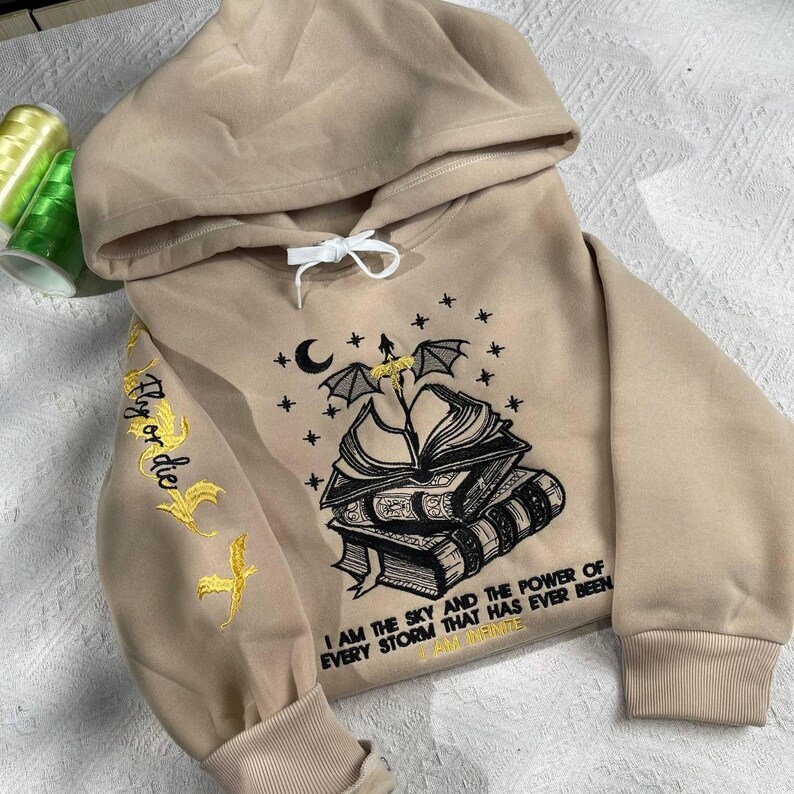 May include: A beige hoodie with a black embroidered design of a stack of books, a crescent moon, and a bat with wings spread. The text "Fly or die" is embroidered in gold on the left sleeve. The text "I am the sky and the power of every storm that has ever been I am infinite" is embroidered in black below the book design.