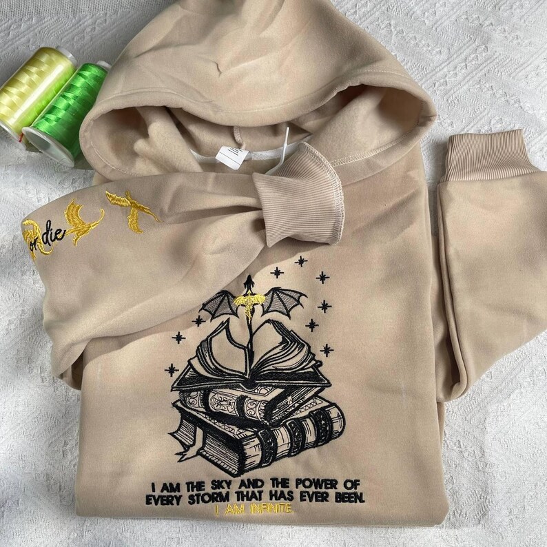May include: A beige hoodie with a black embroidered design of a book with wings and the text "I am the sky and the power of every storm that has ever been. I am infinite". The hoodie has gold embroidery on the sleeve.