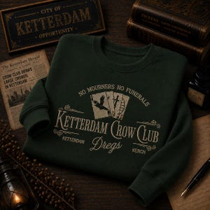 May include: A dark forest green crewneck sweatshirt featuring a graphic print in cream and red. The design includes three playing cards, a crow silhouette, and the text "NO MOURNERS NO FUNERALS", "KETTERDAM CROW CLUB", "DREGS", "KETTERDAM", and "KERCH".