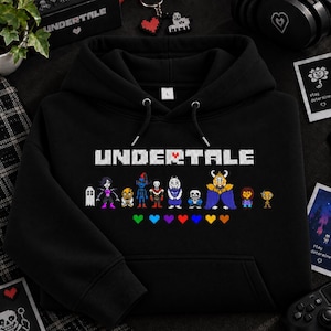 May include: A black hooded sweatshirt featuring the white text UNDERTALE with a red heart replacing the letter A. Below the text are pixelated character illustrations and a row of eight colourful pixelated hearts in various shades.