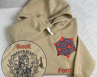 May include: A tan hoodie with a black and white embroidered design on the back that says "Shadowheart Baldur's Gate" and a red and gray embroidered design on the front that looks like a dice.
