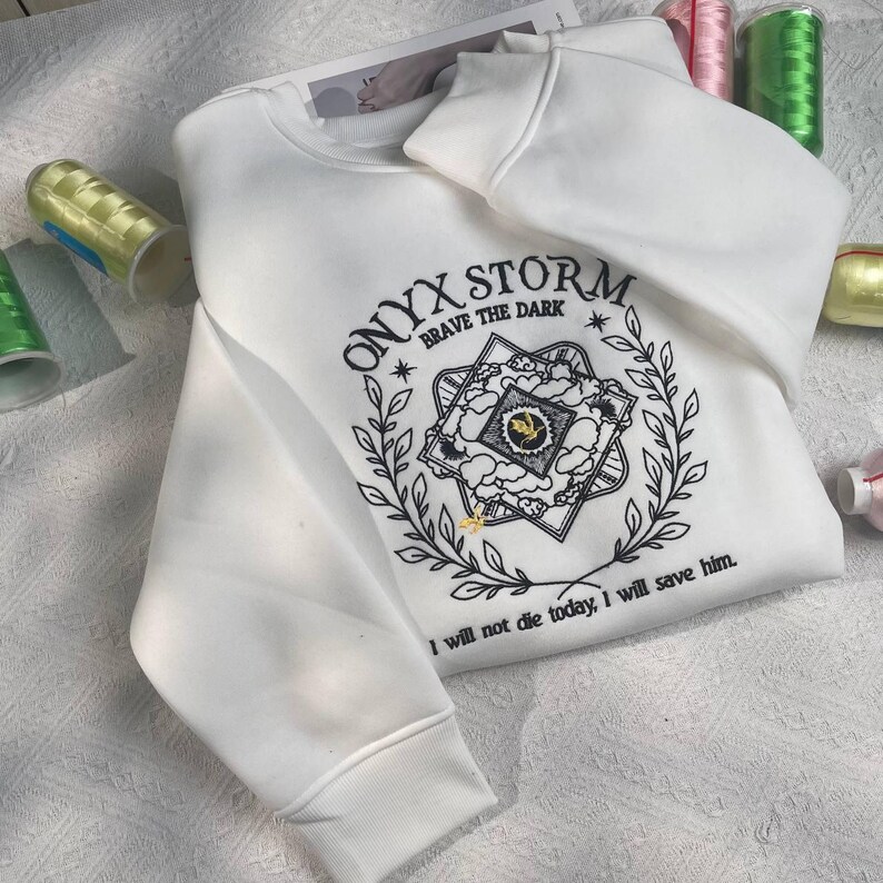 Może przedstawiać: Biała bluza z czarnym haftowanym tekstem, kt&oacute;ry brzmi "ONYX STORM. BRAVE THE DARK" i grafiką gwiazdy z chmurami i liśćmi. Tekst pod grafiką brzmi "Nie umrę dzisiaj, uratuję go."