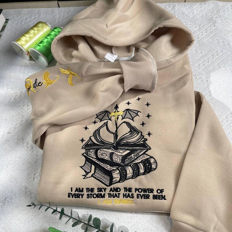 May include: Beige hooded sweatshirt with black embroidered design of a stack of books with a winged creature and stars. The text "I am the sky and the power of every storm that has ever been. I am infinite" is embroidered below the design.