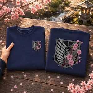 Könnte beinhalten: Zwei marineblaue Sweatshirts mit Rundhalsausschnitt und einem gestickten Emblem aus Schild, Flügeln und Schwert, verziert mit rosa Kirschblütenzweigen. Ein Sweatshirt zeigt ein kleines Emblem auf der Brust, das andere eine größere Version.
