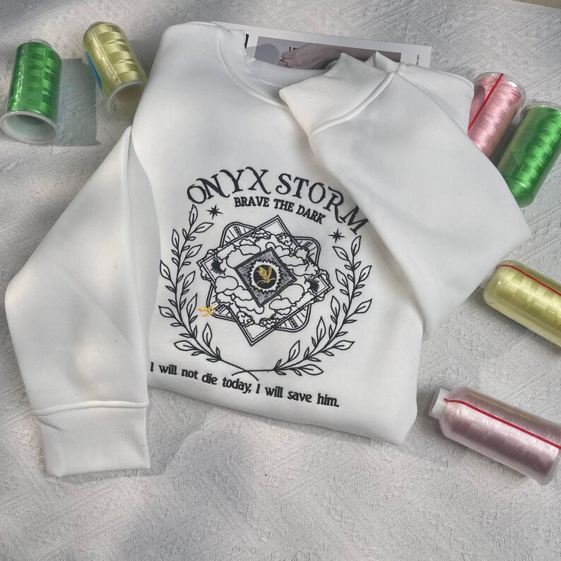 Może przedstawiać: Biała bluza z czarnym nadrukiem graficznym gwiazdy otoczonej kwiatowym wzorem. Tekst "ONYX STORM BRAVE THE DARK" znajduje się nad gwiazdą, a tekst "I will not die today, I will save him." pod gwiazdą.