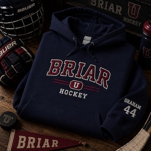 Puede incluir: Una sudadera con capucha azul marino que presenta el texto bordado BRIAR U HOCKEY en granate y blanco en el pecho. La manga izquierda muestra el nombre GRAHAM y el número 44 en bordado blanco. La sudadera tiene un bolsillo canguro frontal.