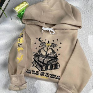 May include: A beige hooded sweatshirt with a black and white embroidered design of a stack of books, a crescent moon, and a bat. The text "Flag or die" is embroidered on the left sleeve. The text "I am the sky and the power of every storm that has ever been. Ana Wente" is embroidered on the front.