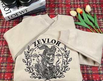 Op de afbeelding: Een beige sweatshirt met een zwarte grafische print van een demonenfiguur met hoorns en een kroon. De grafiek is omringd door een krans van bladeren en de tekst "Zevlor Baldur's Gate".