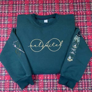 Könnte beinhalten: Dunkelgrünes Sweatshirt mit dem Wort "unlimited" in goldener Schrift auf der Brust. Der linke Ärmel zeigt ein goldenes Zauberstab-Design, der rechte Ärmel eine himmlische Grafik mit Uhren und Sternen.