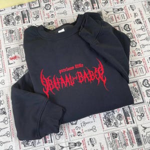 Puede incluir: Sudadera negra con el texto rojo "precious little BHAAL-BABER" bordado en el pecho. La sudadera es de cuello redondo y manga larga. El texto está en una fuente gótica.