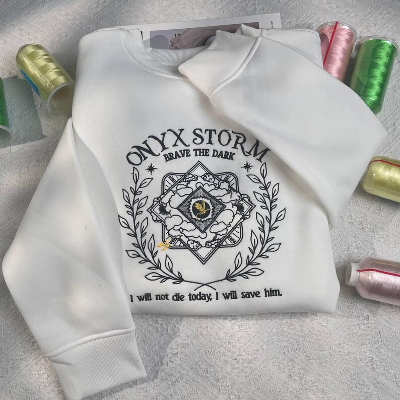 Może przedstawiać: Biała bluza z czarnym haftem z tekstem "ONYX STORM * BRAVE THE DARK *" i grafiką stylizowanej chmury z ptakiem w środku. Na bluzie jest r&oacute;wnież wyszyty tekst "Nie umrę dzisiaj, uratuję go."