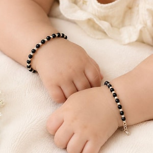 Puede incluir: Dos pulseras a juego que presentan una alternancia de pequeñas cuentas negras y cuentas metálicas de tono plateado. Cada pulsera está diseñada con una delicada cadena de extensión y un pequeño cierre, colocadas en las muñecas de un niño pequeño.