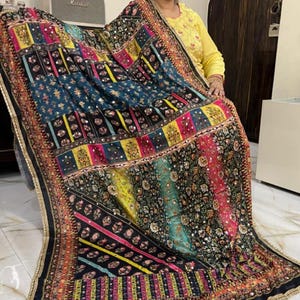 May include: A black and multi-coloured patterned shawl with sequins and embroidery. The shawl has a geometric design with stripes and floral patterns. The shawl is trimmed with a gold and black border.