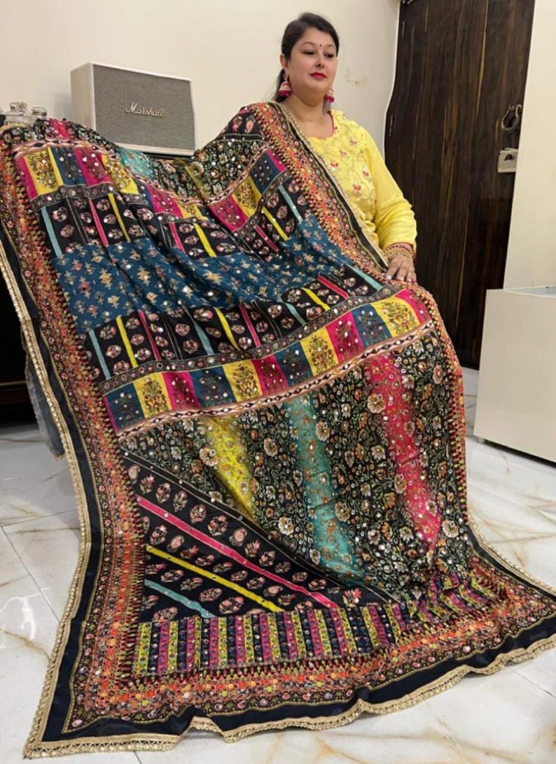 May include: A black and multi-coloured patterned shawl with sequins and embroidery. The shawl has a geometric design with stripes and floral patterns. The shawl is trimmed with a gold and black border.