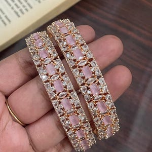 Può includere: Due braccialetti d'oro con pietre rosa e bianche. I braccialetti sono decorati con un motivo ripetitivo di pietre rosa rettangolari e pietre bianche più piccole.