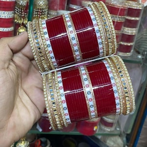 Può includere: Un paio di braccialetti rossi e dorati con dettagli bianchi e dorati. I braccialetti presentano un motivo ripetuto di cerchi rossi, bianchi e blu. Sono decorati con piccoli specchi e finiture dorate. Gioielli tradizionali indiani.