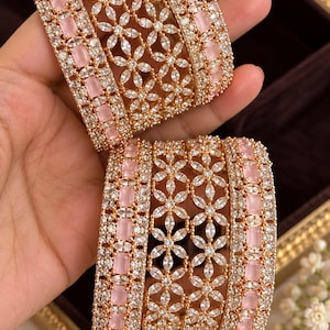 May include: A set of two gold-toned bangles featuring a central band of light pink rectangular stones flanked by rows of clear marquise-cut stones arranged in a floral pattern, with additional clear stones lining the edges of the metal bands.