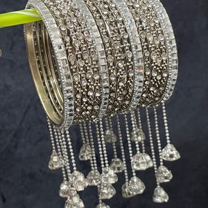 May include: A stack of silver bangles with clear, sparkling rhinestones and dangling bell charms. The bangles are arranged on a green stick against a dark background. The bangles are a mix of solid bands and bands with intricate rhinestone detailing.