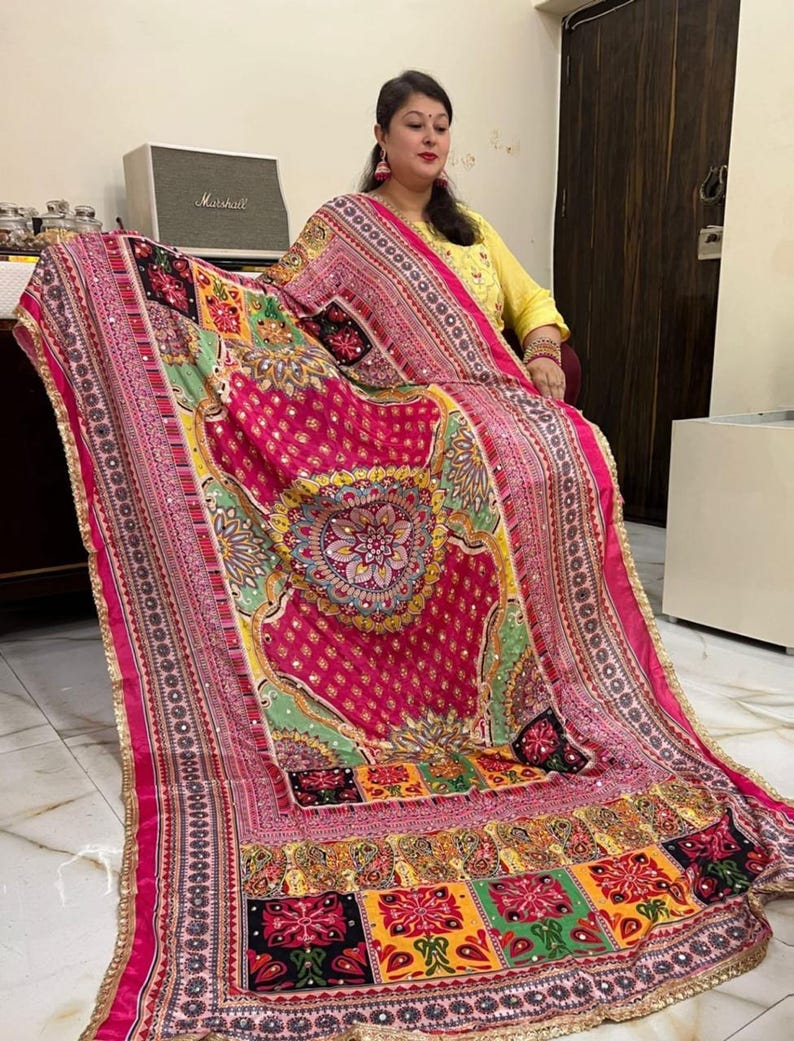 May include: A pink and green patterned shawl with gold trim and sequins. The shawl features a large circular design in the centre and a patchwork design at the bottom.