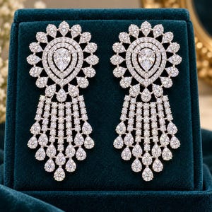 May include: A pair of silver-toned drop earrings featuring a central pear-shaped stone surrounded by multiple layers of small, clear, round and pear-shaped stones arranged in a sunburst pattern with a cascading fringe of dangling stones.