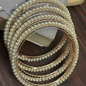 Peut inclure: Ensemble de quatre bracelets dorés avec des accents de perles blanches. Les bracelets sont empilés les uns sur les autres.