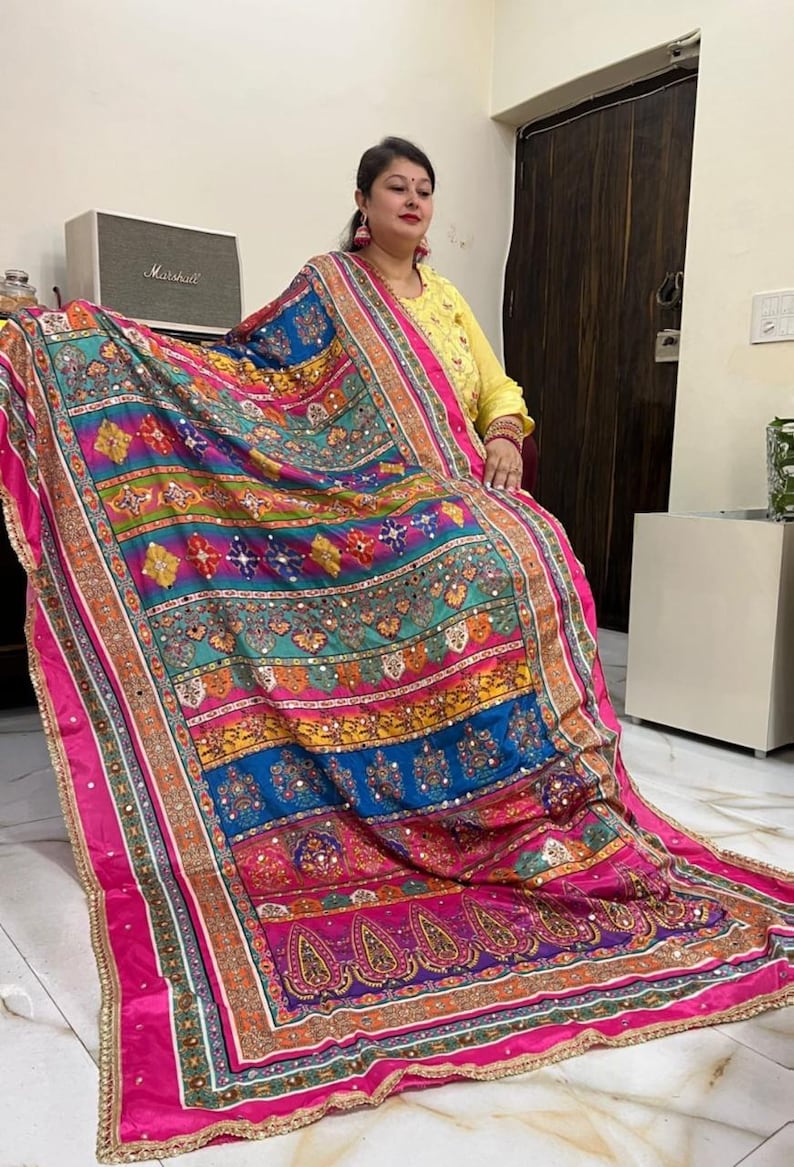 May include: A colourful, multi-patterned shawl with a pink border and gold accents. The shawl is made of a soft, flowing fabric and features intricate embroidery and mirror work.