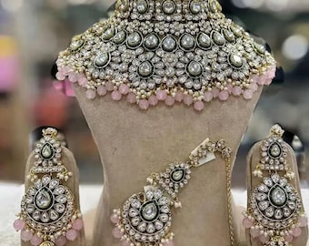 May include: A traditional Indian bridal jewelry set featuring a large, ornate choker necklace with green and white stones, pink beads, and a matching pair of earrings and a maang tikka.