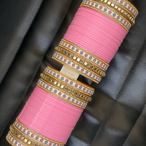 Può includere: Due braccialetti rosa con dettagli dorati, bianchi e a specchio. I braccialetti sono impilati e tenuti su un supporto bianco. Il design include piccoli accenti rossi e blu, creando un aspetto festoso. Accessorio tradizionale.