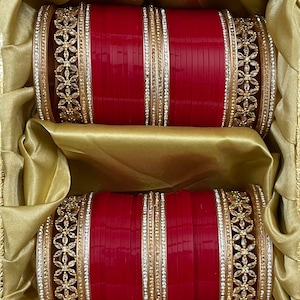 Può includere: Un paio di braccialetti rossi con dettagli dorati e argentati, presentati in una scatola foderata in raso dorato. I braccialetti presentano intricati disegni floreali e di strass, creando un aspetto lussuoso. La scatola aggiunge alla presentazione.