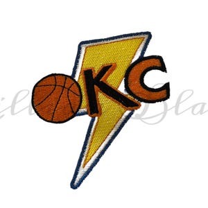 Puede incluir: Parche bordado con una pelota de baloncesto y las letras "KC" en naranja y negro, sobre un rayo amarillo con borde azul. El diseño está sobre un fondo blanco.