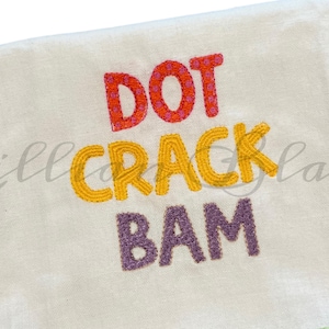 May include: A white towel with the embroidered words "DOT CRACK BAM" in orange, yellow, and purple. The towel has a soft, textured appearance.
