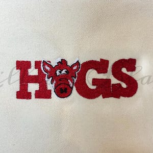 May include: Cream-colored fabric with the word "HOGS" in red, featuring a cartoon Razorback hog in the center. The design is likely for a sports fan or supporter of the University of Arkansas.