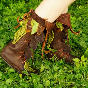 May include: Brown leather boots with green and brown leaf-shaped embellishments. The boots are worn by a person standing in a grassy area.
