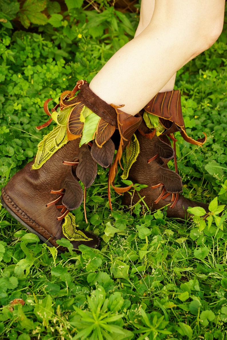 May include: Brown leather boots with green and brown leaf-shaped embellishments. The boots are worn by a person standing in a grassy area.
