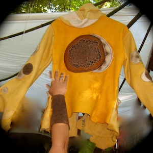 Puede incluir: Una chaqueta amarilla con capucha con un parche de cuero marrón que presenta un diseño geométrico. La chaqueta tiene una capucha blanca y está decorada con varios parches de cuero marrón.