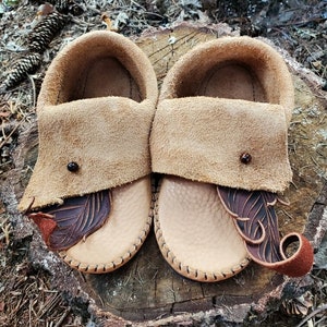 May include: A pair of brown leather moccasins with decorative stitching and a feather design. The moccasins are sitting on a tree stump.