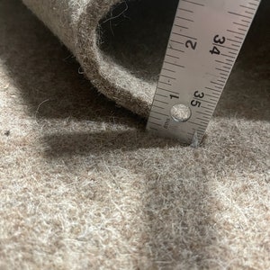 K&ouml;nnte beinhalten: Nahaufnahme eines beigefarbenen Filzstoffs mit einem Lineal, das 84 cm misst. Der Stoff ist weich und texturiert, mit einem leichten Glanz.