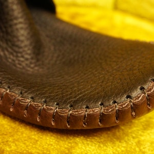 May include: Close-up of a brown leather moccasin with a brown stitched seam around the edge.