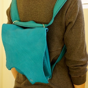 Peut inclure: Un sac à dos en cuir turquoise avec des coutures noires sur les bretelles et un détail d'anneau doré. Le sac à dos est porté sur une chemise grise à manches longues et un jean bleu.