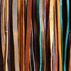 Op de afbeelding: Een assortiment van leren koorden in verschillende kleuren en breedtes, waaronder bruin, beige, blauw, groen en paars.