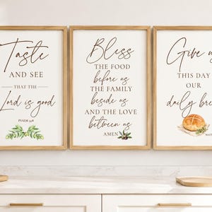 May include: A set of three rectangular wall art prints in light wood frames. Each print features white backgrounds with black script and serif text. The first reads Taste and see that the Lord is good, the second Bliss the food before us, and the third Give us this day our daily bread with a small illustration of a loaf of bread.