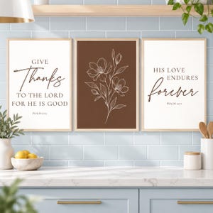 May include: A set of three framed wall art prints. The left print has a white background with dark brown text reading "GIVE Thanks TO THE LORD FOR HE IS GOOD PSALM 107:1". The centre print features a white line drawing of flowers on a brown background. The right print has a white background with dark brown text reading "HIS LOVE ENDURES forever PSALM 107:1".