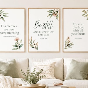 May include: Three vertical wall art prints in light wood frames featuring white backgrounds with botanical illustrations of green leaves and pink flowers. The text reads: His mercies are new every morning, Be still and know that I am God, and Trust in the Lord with all your heart.