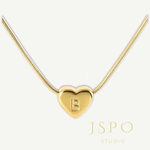 May include: A gold-colored necklace with a heart-shaped pendant. The pendant features the letter "B" and is suspended from a delicate chain. The image also includes the text "JSPO STUDIO".