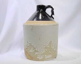 May include: A vintage stoneware jug with a dark brown glazed top and handle. The body of the jug is a creamy white with a textured, aged appearance. The jug has a classic, rustic design, suitable for decorative use.